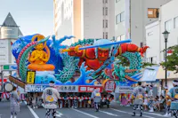 A vibrant parade float featuring a fierce red warrior with a blue dragon and a golden Buddha statue is paraded down a city street by people in traditional Japanese festival attire. Spectators watch from the sidewalks.