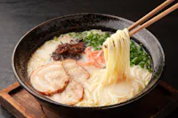 A bowl of ramen with sliced pork, green onions, mushrooms, and pickled ginger in a creamy broth, with chopsticks lifting noodles from the bowl.