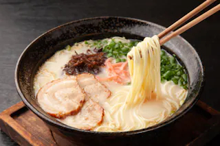 A bowl of ramen with sliced pork, green onions, mushrooms, and pickled ginger in a creamy broth, with chopsticks lifting noodles from the bowl.