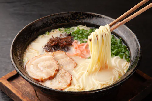 A bowl of ramen with sliced pork, green onions, mushrooms, and pickled ginger in a creamy broth, with chopsticks lifting noodles from the bowl.
