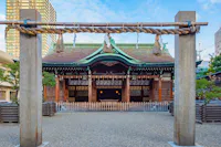 A traditional Japanese Shinto shrine with a decorative torii gate and shimenawa rope in the foreground, wooden offering plaques, and a green copper roof, surrounded by urban buildings.