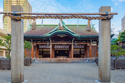 A traditional Japanese Shinto shrine with a decorative torii gate and shimenawa rope in the foreground, wooden offering plaques, and a green copper roof, surrounded by urban buildings.