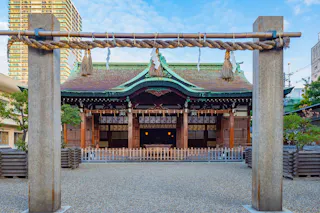 A traditional Japanese Shinto shrine with a decorative torii gate and shimenawa rope in the foreground, wooden offering plaques, and a green copper roof, surrounded by urban buildings.