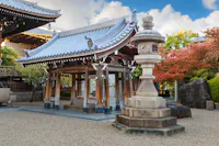 A traditional Japanese temple with intricate wooden architecture and a stone lantern. The courtyard has gravel, and nearby trees display green and red foliage under a partly cloudy sky.