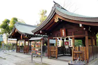 A Japanese Shinto shrine with wooden structures, traditional curved roofs, lanterns, and ema plaques for wishes hanging on a board. The area is peaceful, surrounded by trees and stone paths.