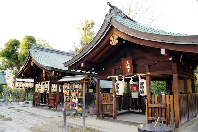 A Japanese Shinto shrine with wooden structures, traditional curved roofs, lanterns, and ema plaques for wishes hanging on a board. The area is peaceful, surrounded by trees and stone paths.