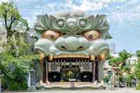 A shrine entrance shaped like a giant, stylized lion or dragon head with wide eyes and large fangs, surrounded by greenery and set against a blue sky with scattered clouds.