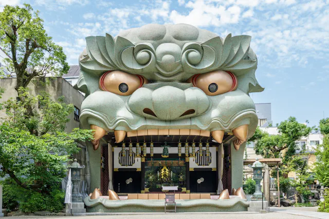 A shrine entrance shaped like a giant, stylized lion or dragon head with wide eyes and large fangs, surrounded by greenery and set against a blue sky with scattered clouds.