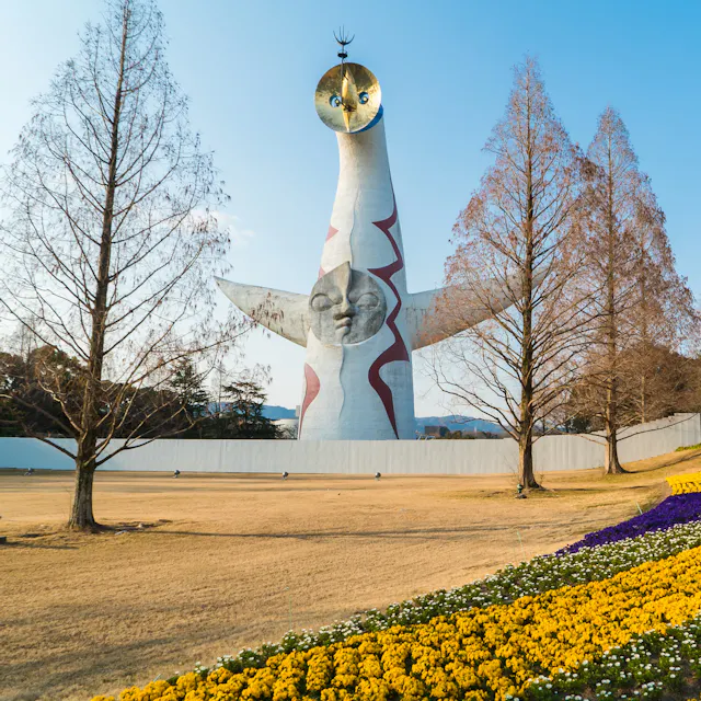 Expo '70 Commemorative Park The image shows the Tower of the Sun, a tall, white sculpture with abstract features, standing in a park with flower beds of yellow and purple blooms and leafless trees under a clear blue sky.