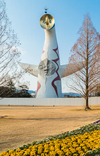 Expo '70 Commemorative Park The image shows the Tower of the Sun, a tall, white sculpture with abstract features, standing in a park with flower beds of yellow and purple blooms and leafless trees under a clear blue sky.