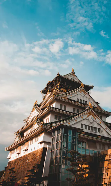 Osaka Castle A traditional Japanese castle with white walls and black-tiled roofs, adorned with gold accents, stands against a blue sky with scattered clouds. Modern glass structures are visible at the castle’s base.