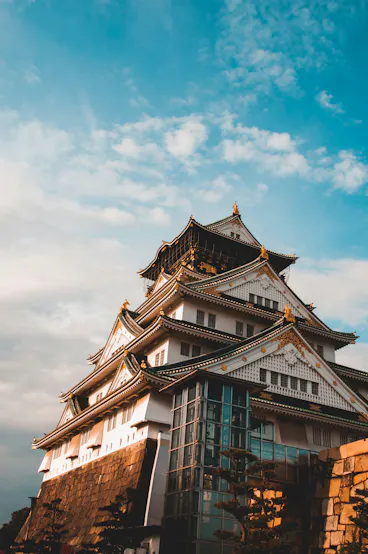 A traditional Japanese castle with white walls and black-tiled roofs, adorned with gold accents, stands against a blue sky with scattered clouds. Modern glass structures are visible at the castle’s base.