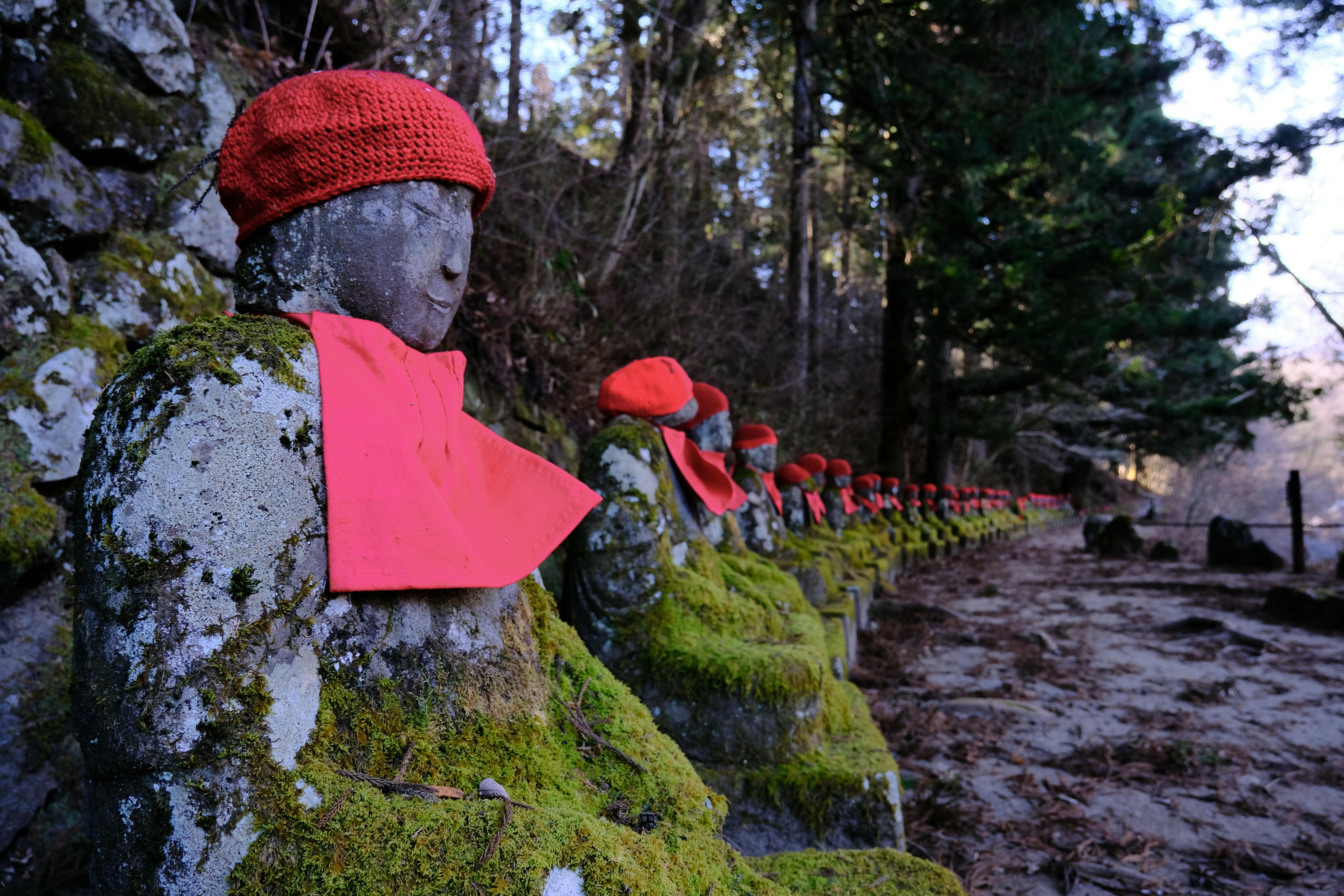 A row of moss-covered stone statues wearing red knitted hats and bibs lines a forest path, with tall trees in the background. The statues appear weathered and serene.