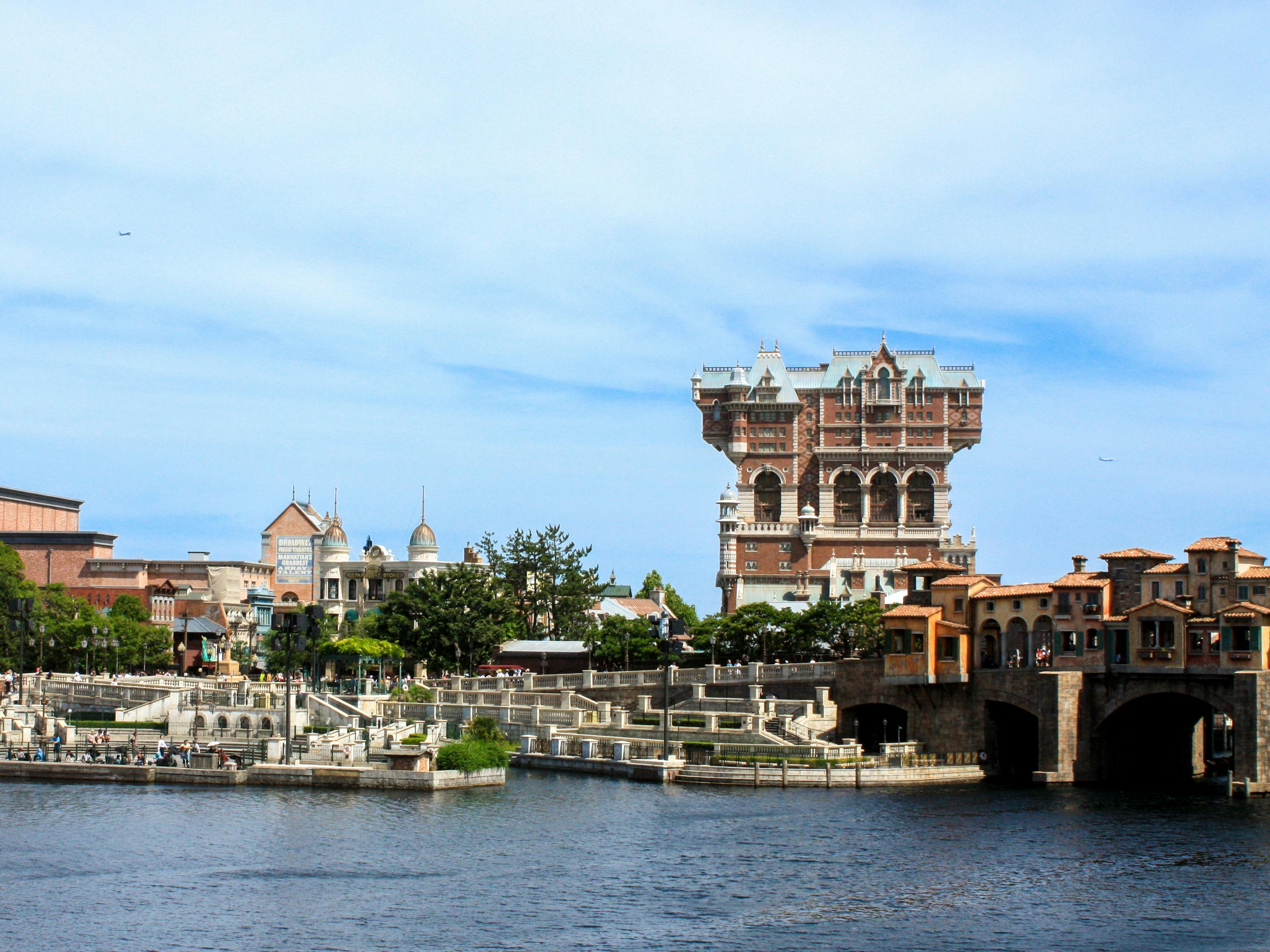 A large waterfront with Mediterranean-style buildings and a tall, ornate, red-brick tower in the background under a blue sky with scattered clouds.