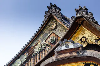 Close-up of ornate, traditional Japanese temple roof with intricate carvings, gold accents, floral patterns, and decorative tiles against a clear blue sky.