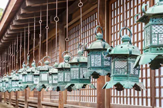 Rows of ornate, green metal lanterns hang from the eaves of a traditional Japanese wooden building with sliding paper doors.