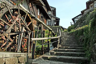 A large wooden waterwheel stands beside a traditional Japanese building, next to a stone staircase bordered by greenery, leading upward under a clear sky.