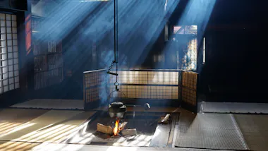 Sunlight streams through windows into a traditional Japanese room with tatami mats, where a kettle hangs over an open hearth fire, surrounded by wooden partitions and light smoke rising from the flames.