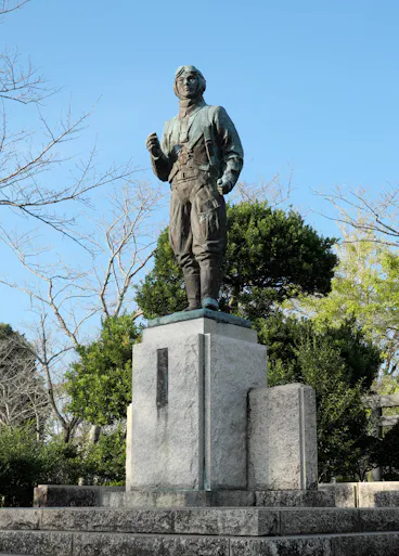 A bronze statue of a man in aviator attire stands on a tall stone pedestal, surrounded by trees and greenery under a clear blue sky.