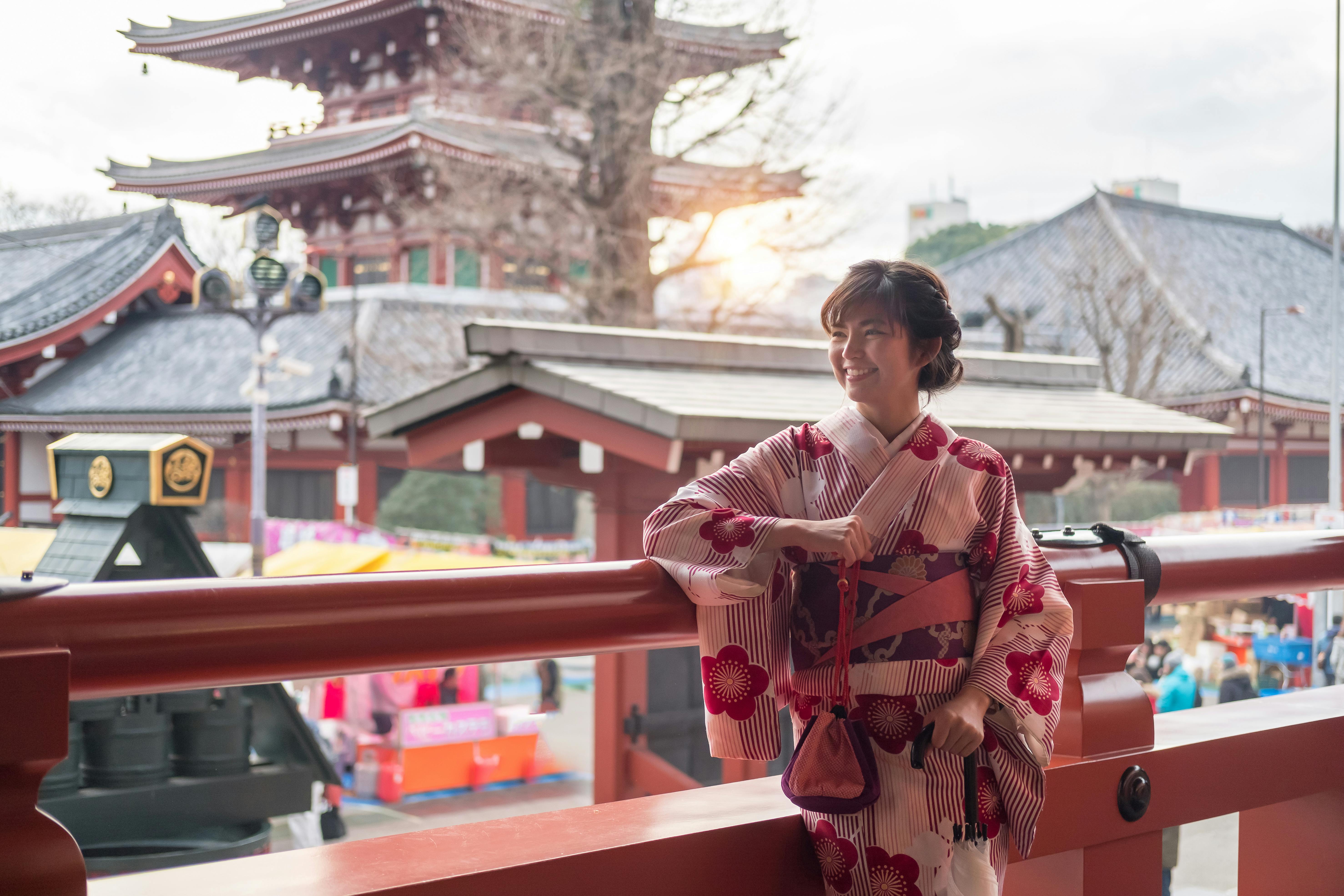 A woman in a pink and white kimono with red patterns stands smiling on a balcony, with traditional Japanese temple buildings and a pagoda visible in the background.