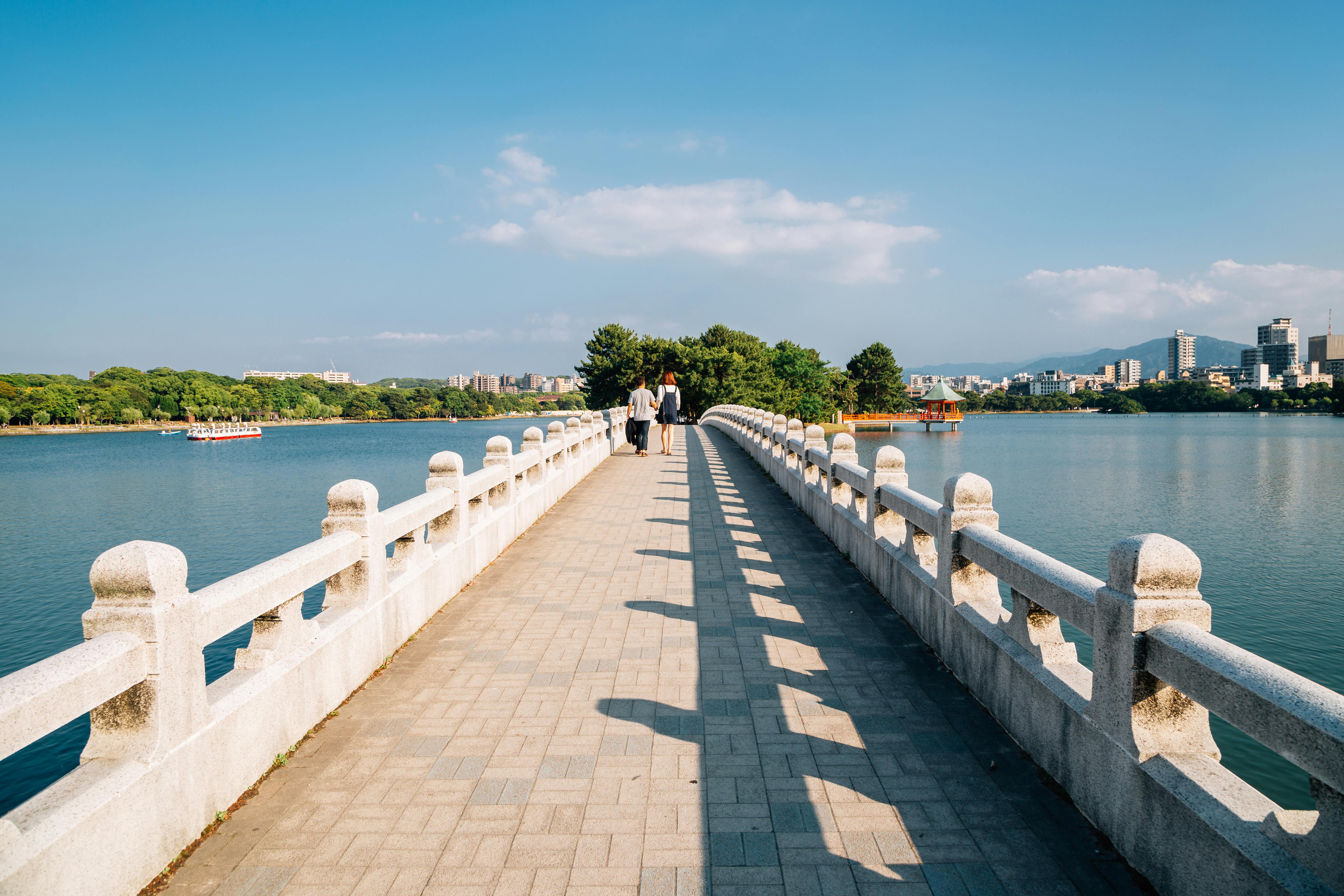 A wide stone bridge with white railings stretches over a calm lake toward trees and distant buildings under a blue sky. A few people walk along the bridge in the sunlight.