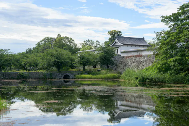 A traditional Japanese castle building with white walls and a tiled roof stands behind a stone wall, surrounded by lush green trees and reflected in a calm, lily-covered pond under a partly cloudy sky.