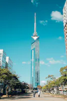 A tall, modern glass skyscraper with a spire stands against a blue sky, surrounded by trees and a few people walking on a paved path in the foreground.