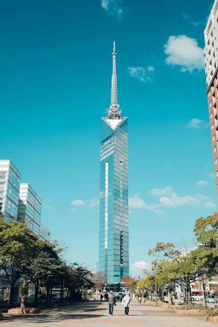 A tall, modern glass skyscraper with a spire stands against a blue sky, surrounded by trees and a few people walking on a paved path in the foreground.