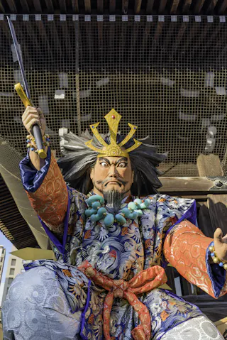 A large, colorful statue of a warrior in ornate traditional Japanese attire and crown, holding a staff and looking stern, on display outdoors under a wooden structure.
