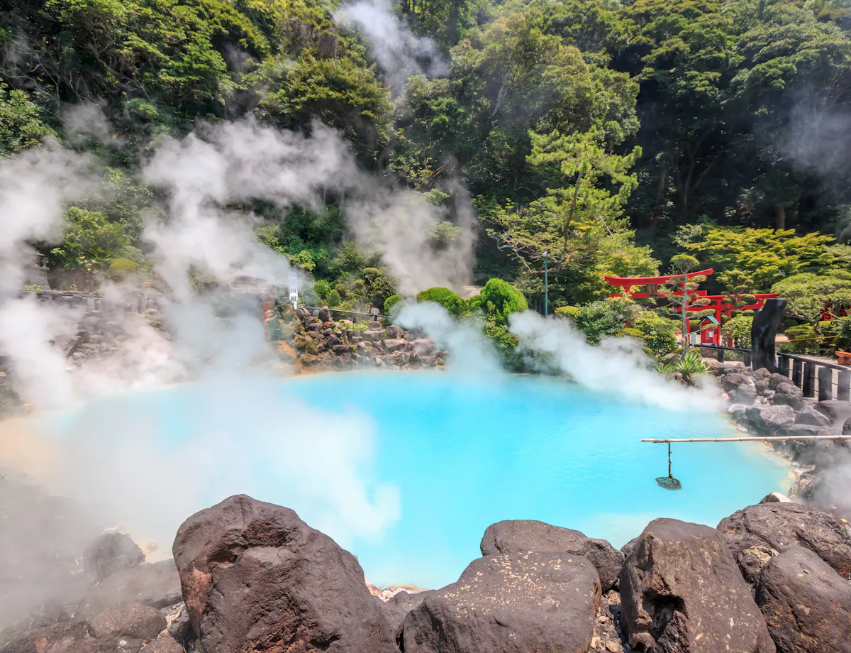 Umijigoku A steaming blue hot spring surrounded by rocks and lush green trees, with red torii gates visible on the right and mist rising from the water.
