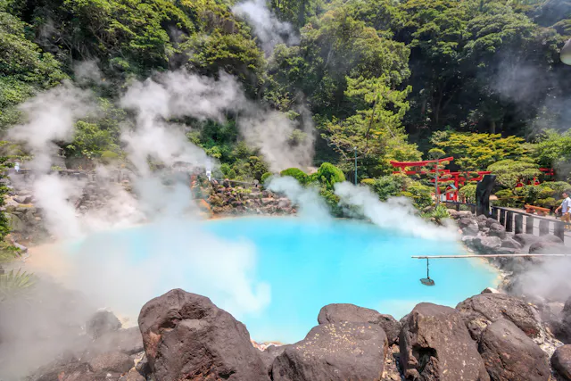 A steaming blue hot spring surrounded by rocks and lush green trees, with red torii gates visible on the right and mist rising from the water.
