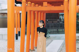 A woman in a black top and tan dress stands smiling among vibrant orange torii gates at a traditional Japanese shrine.