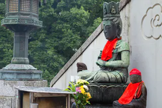 A large and a small stone Buddha statue, both wearing bright red cloths, sit beside a wall with flowers placed in front. Lush green trees and an ornate lantern are visible in the background.