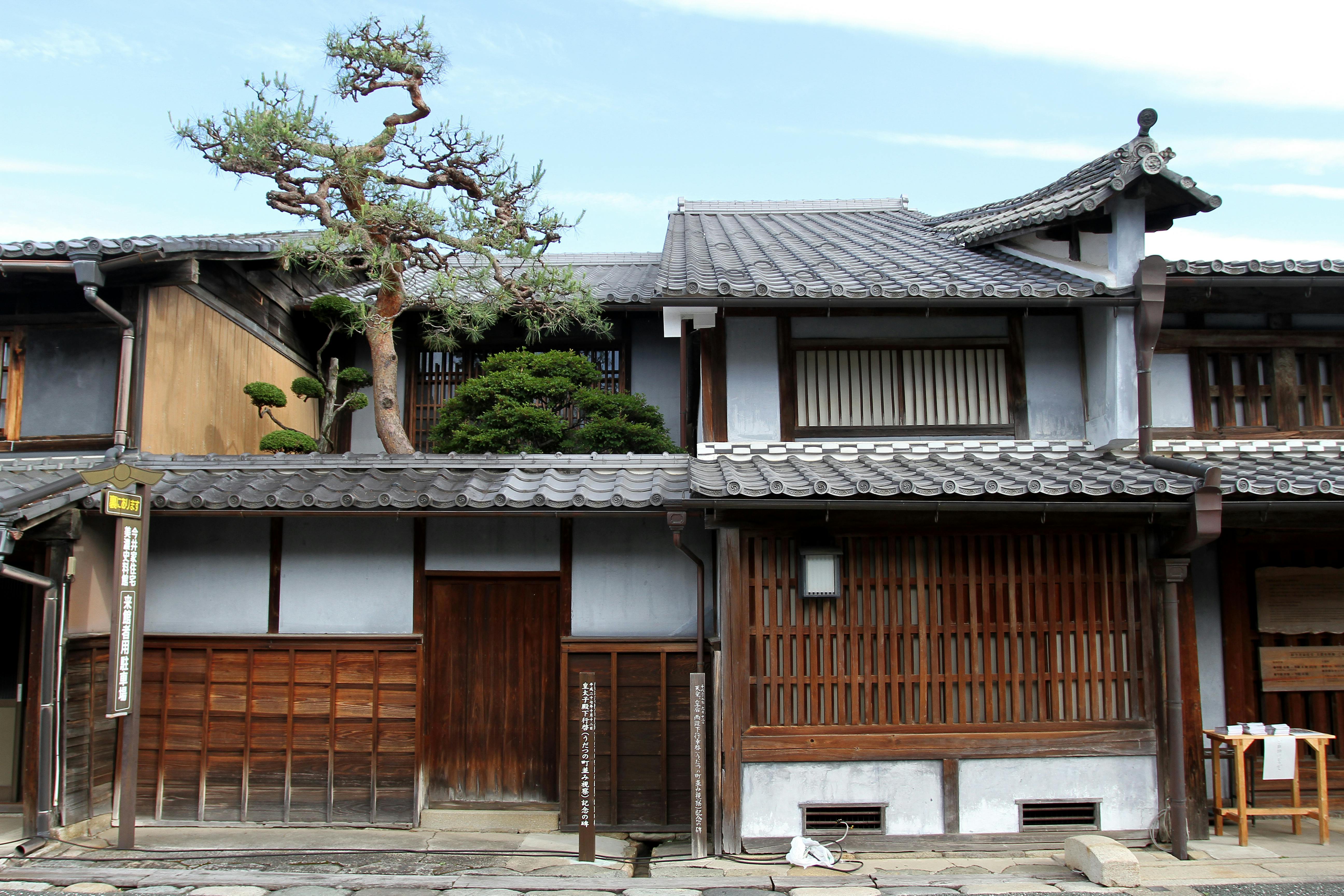A traditional Japanese wooden house with sliding doors, tiled roof, and a manicured tree visible behind the fence, set along a stone-paved street under a blue sky.