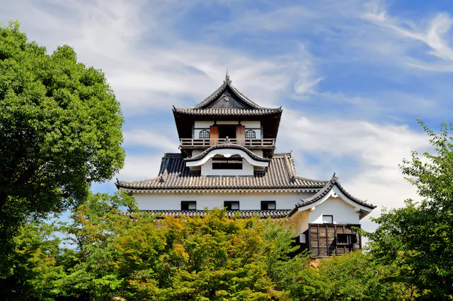 A traditional Japanese castle with tiered, tiled roofs and ornate details, partially obscured by lush green trees under a bright blue sky with scattered clouds. The castle is a prominent historical structure surrounded by greenery.