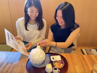 Two women smile while sitting at a table with a large bowl of shaved ice dessert topped with fruit and syrup in front of them. One woman holds a menu, and the other reaches to add a topping.