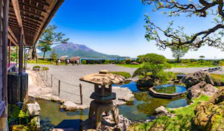 Traditional Japanese garden with a stone lantern, pond, manicured trees, and gravel courtyard; people walk in the distance with a scenic view of a mountain under a clear blue sky.