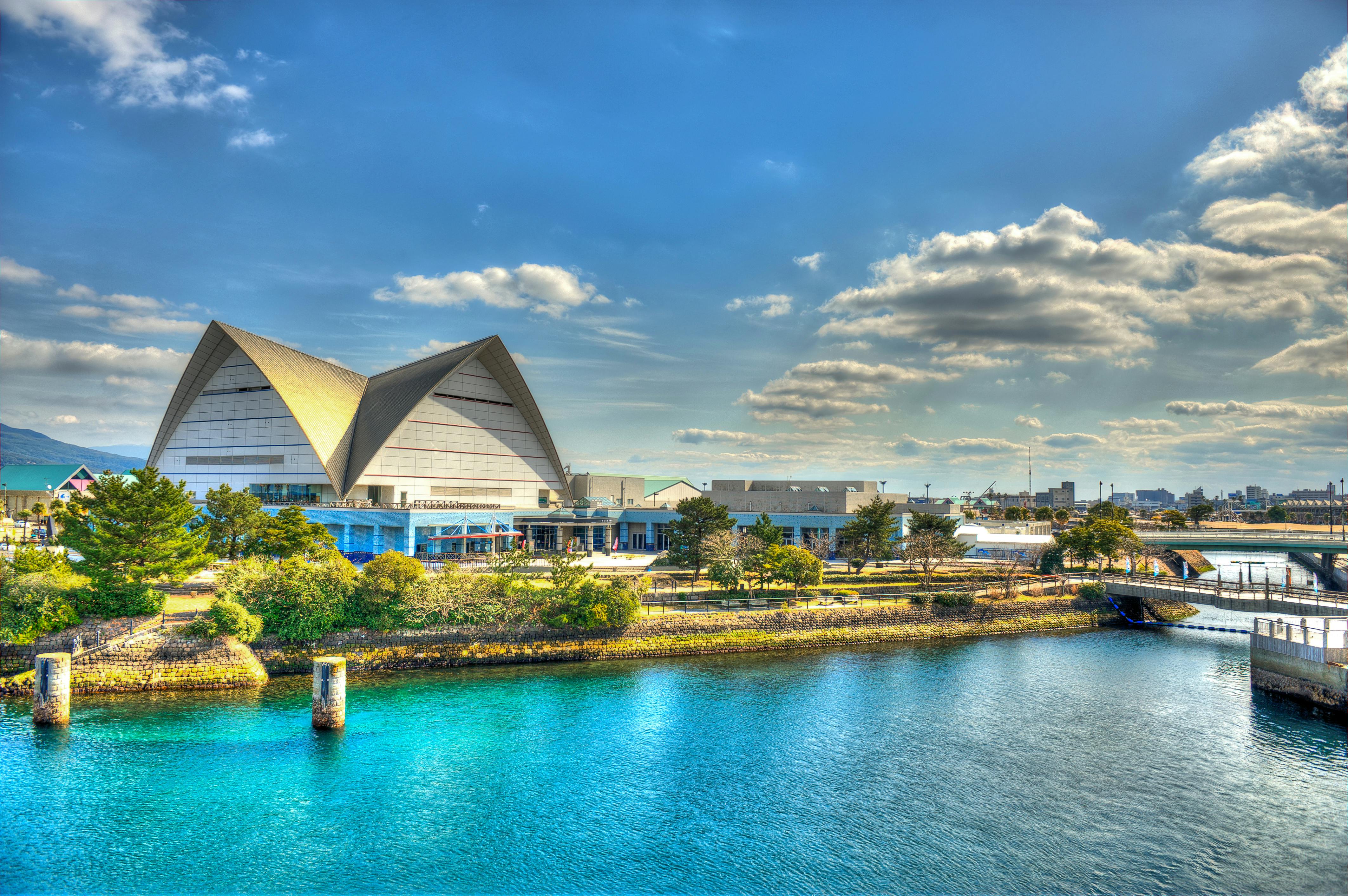 A modern building with a distinctive angular roof sits by clear blue water and greenery under a partly cloudy sky, with a bridge and cityscape visible in the background.