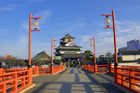 A traditional Japanese castle stands at the end of a red bridge lined with lanterns under a clear blue sky, surrounded by trees and buildings.