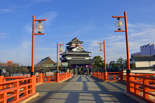 A traditional Japanese castle stands at the end of a red bridge lined with lanterns under a clear blue sky, surrounded by trees and buildings.