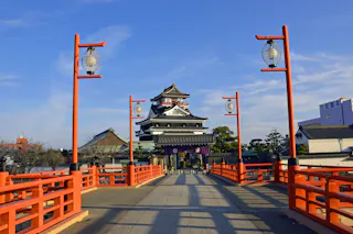 A traditional Japanese castle stands at the end of a red bridge lined with lanterns under a clear blue sky, surrounded by trees and buildings.