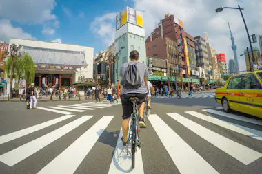 A person wearing a striped shirt and backpack rides a bicycle across a busy crosswalk in an urban area with tall buildings, a temple, many pedestrians, and a yellow taxi under a blue sky.