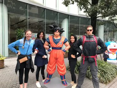 Four people pose playfully with hands on hips beside a life-sized statue of Goku from Dragon Ball Z on a city sidewalk, with glass buildings and trees in the background.