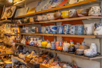 Shelves display a variety of colorful ceramic cups, bowls, teapots, plates, and dishes in a shop, with intricate patterns and traditional Japanese designs. The scene is warm and densely packed with pottery items.