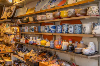 Shelves display a variety of colorful ceramic cups, bowls, teapots, plates, and dishes in a shop, with intricate patterns and traditional Japanese designs. The scene is warm and densely packed with pottery items.