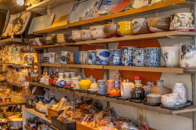 Shelves display a variety of colorful ceramic cups, bowls, teapots, plates, and dishes in a shop, with intricate patterns and traditional Japanese designs. The scene is warm and densely packed with pottery items.