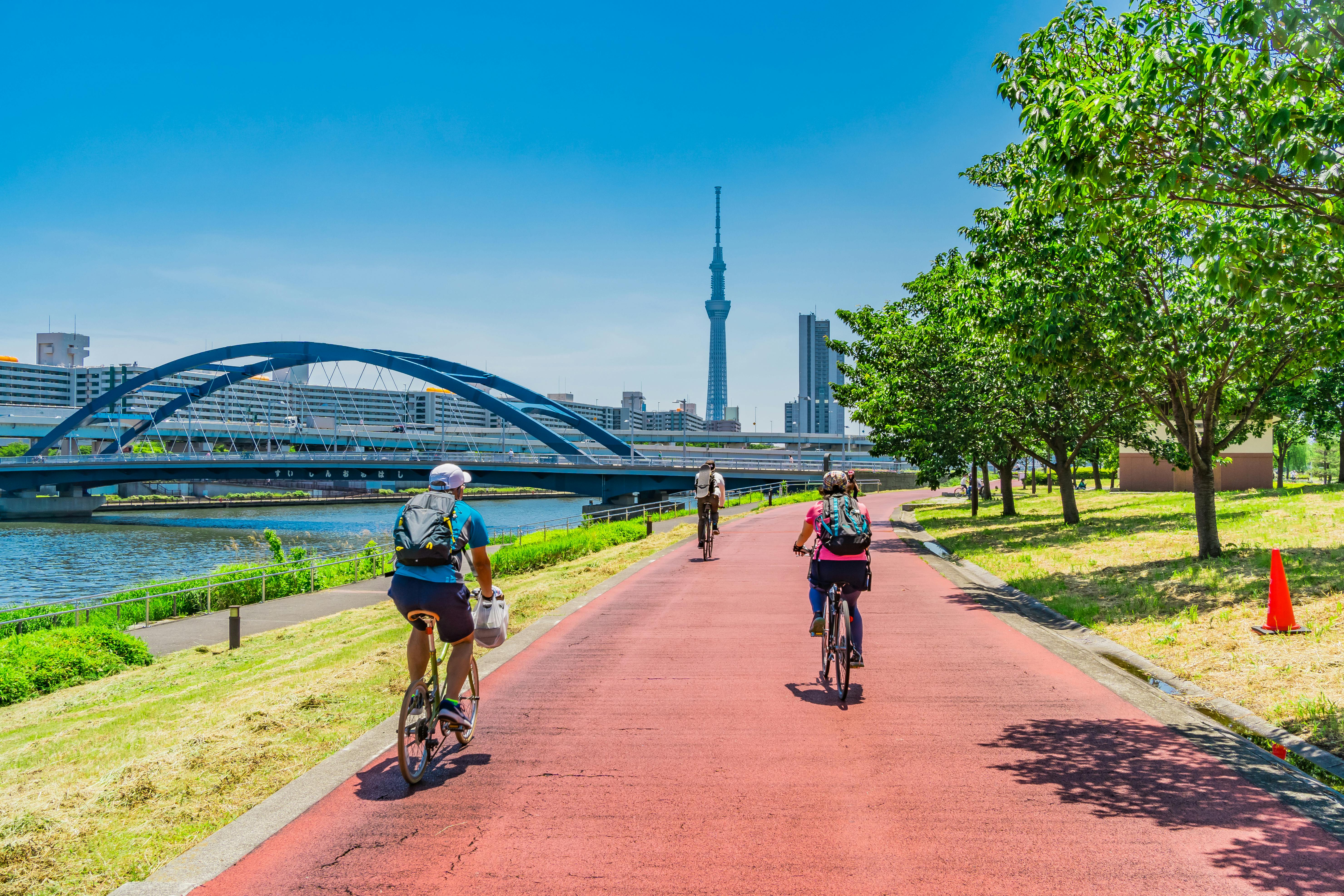 People ride bicycles on a red path along a river with green trees and grass. A blue bridge, city buildings, and the Tokyo Skytree tower are visible under a clear blue sky.