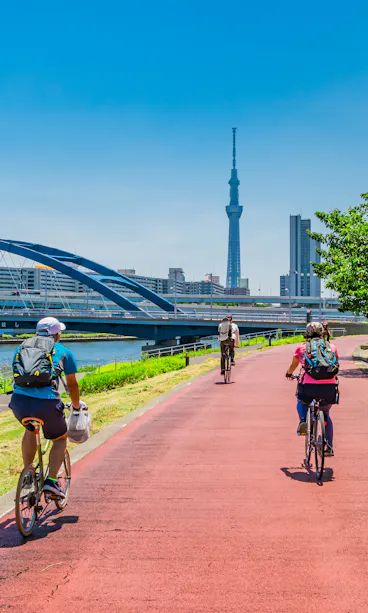 Cycling in Tokyo People ride bicycles on a red path along a river with green trees and grass. A blue bridge, city buildings, and the Tokyo Skytree tower are visible under a clear blue sky.