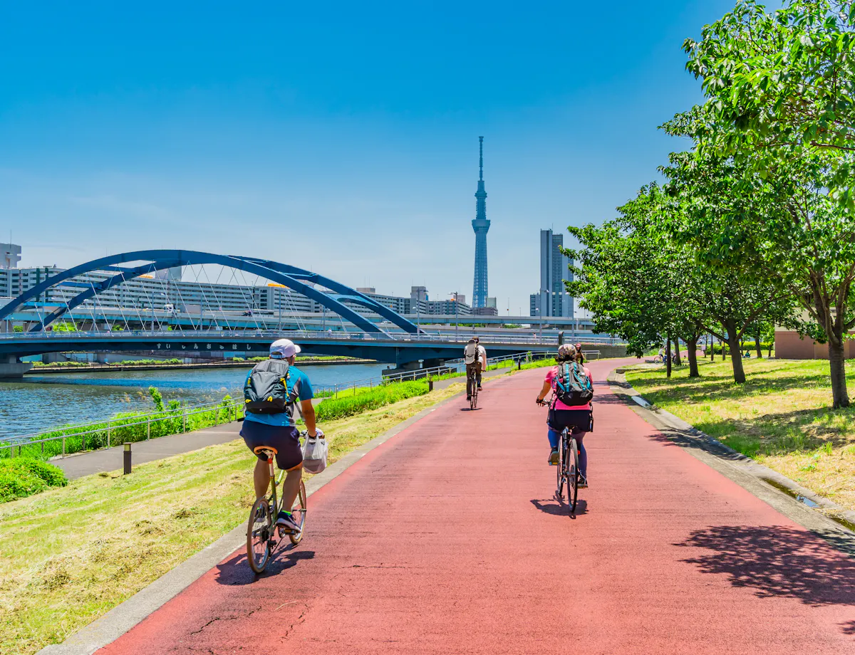 Cycling in Tokyo People ride bicycles on a red path along a river with green trees and grass. A blue bridge, city buildings, and the Tokyo Skytree tower are visible under a clear blue sky.