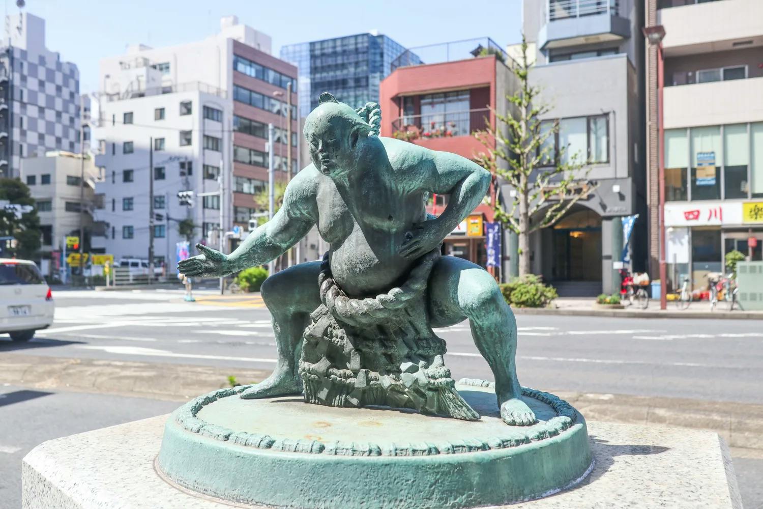 A bronze statue of a sumo wrestler in a traditional stance stands on a circular pedestal, with city buildings, trees, and a street visible in the background on a sunny day.